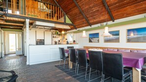 Un vaste espace de salle à manger avec un haut plafond et des poutres apparentes, une cuisine dotée d’un îlot central et une vue sur l’extérieur.
