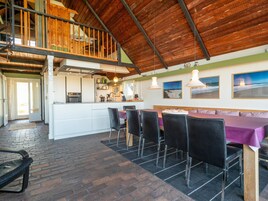 A spacious dining area with a high ceiling and exposed wooden beams, a kitchen with a central island, and a view of the outdoors.