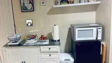 Ett litet, praktiskt kök med mikrovågsugn, ett litet kylskåp, en papperskorg, en bänkskiva med en skål med frukt och en hylla med köksredskap.