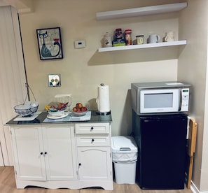 Ett litet, praktiskt kök med mikrovågsugn, ett litet kylskåp, en papperskorg, en bänkskiva med en skål med frukt och en hylla med köksredskap.