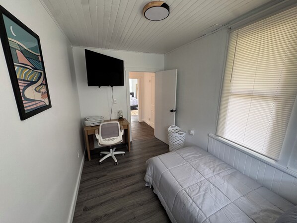 Ein Schlafzimmer mit Bett, Schreibtisch mit Stuhl, Wand-Fernseher, Fenster mit Jalousien und einer Tür zu einem weiteren Raum.