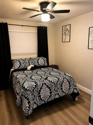 Une chambre à coucher avec un lit, un ventilateur de plafond, une fenêtre avec des stores et deux œuvres d’art encadrées accrochées au mur.