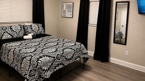 A bedroom with a bed, black curtains, a framed picture, a mirror, and a wall-mounted TV. - Signature Stay 5 Minutes From Hard Rock Stadium (Miami Gardens)