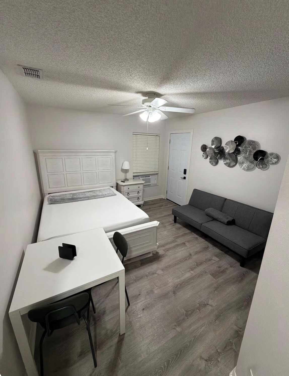 A bedroom with a bed, a desk, a chair, a gray sofa, a ceiling fan, a wall-mounted light, and a decorative wall piece.