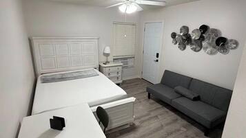 A bedroom with a bed, a desk, a chair, a gray sofa, a ceiling fan, a wall-mounted light, and a decorative wall piece.
