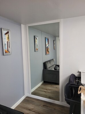 Ein Zimmer mit grauer Wand, Holzboden, einem grauen Sofa mit Kissen und einem Spiegel, der das Zimmer reflektiert.
