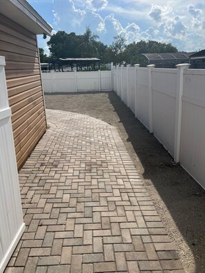 Ein gepflasterter Patio-Bereich mit weißem Zaun, einem Holzgebäude und einer überdachten Struktur im Hintergrund.