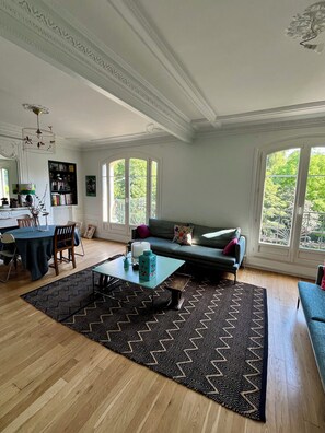 A spacious living room with a dining area, large windows, and a decorative ceiling. - Haussmann Charm · Facing the Woods · 15 min to Central Paris/Disney (Vincennes)