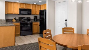 A compact kitchen with wooden cabinets, a black refrigerator, a microwave, and an oven. There is a dining table with four chairs.