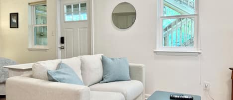 A living room with a sofa, a round mirror, a small table, and a window with a view of a building.