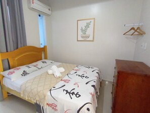 Ein Schlafzimmer mit einem Bett, einem Holzkopfteil, einem Nachttisch, einem Schminktisch und einem Bild in einem Rahmen an der Wand. Ein Fenster mit Vorhängen ist ebenfalls zu sehen.