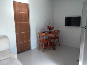 Une pièce avec une porte en bois, une table de salle à manger ronde entourée de chaises, une petite plante en pot et un téléviseur à écran plat fixé au mur.