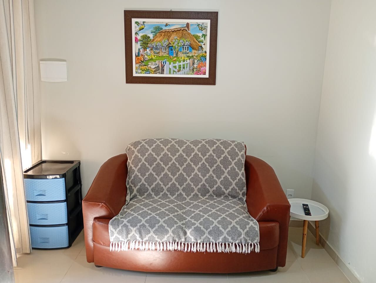A room with a brown leather sofa, a small round table, a framed painting on the wall, and a set of blue drawers.