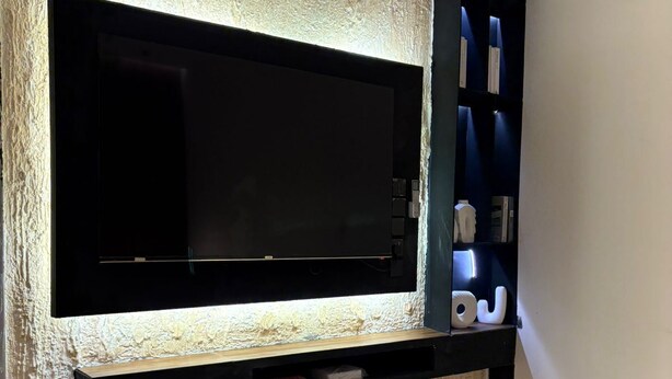 A living room with a large flat-screen TV mounted on a textured wall, a glass coffee table, and a bookshelf with decorative items. - OrbitNet-Al Khatim (Madina)