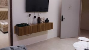 A modern living room with a flat-screen TV mounted on the wall, a wooden shelf with decorative items, a grey sofa, a round coffee table, and a patterned rug. - OrbitNet-Al Khatim (Madina)