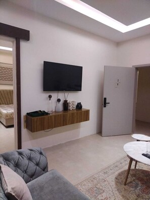 Ein modernes Wohnzimmer mit einem an der Wand befestigten Flachbildfernseher, einem Holzregal mit Dekoration, einem grauen Sofa, einem runden Couchtisch und einem gemusterten Teppich.