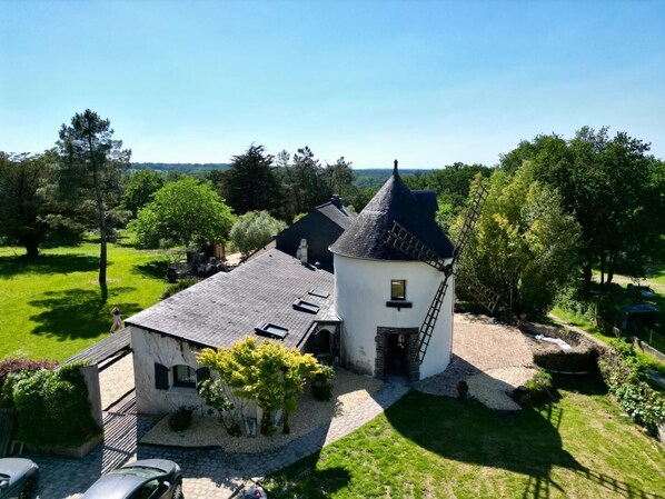 A white building with a black roof and a windmill, surrounded by greenery and trees.