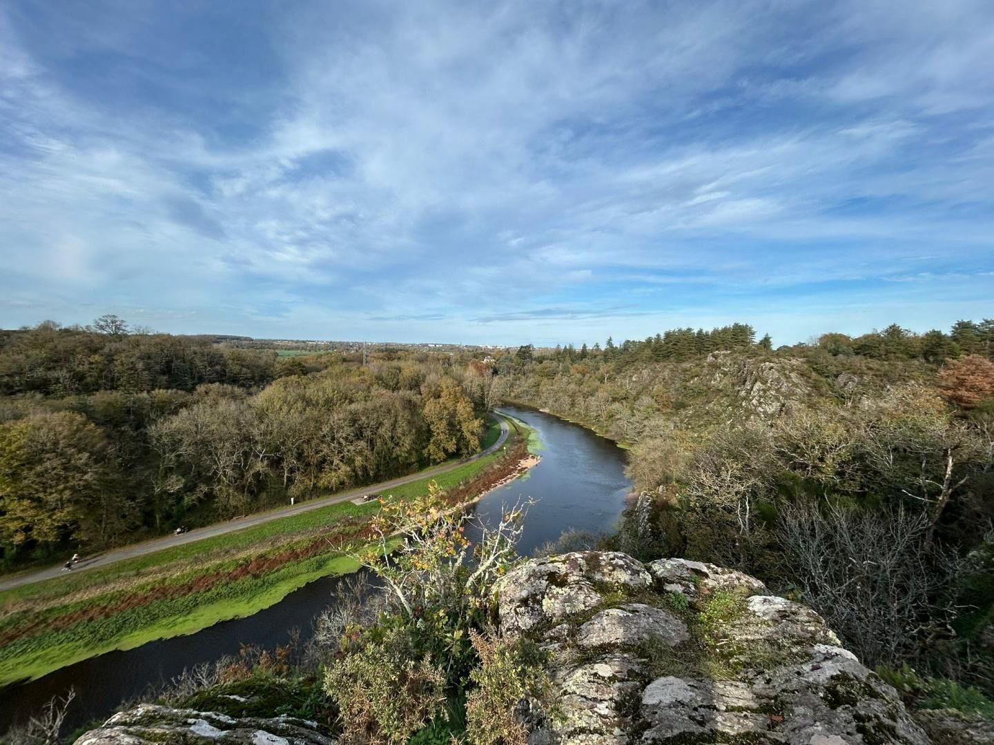 A river with a road alongside it, surrounded by trees and a stone wall.
