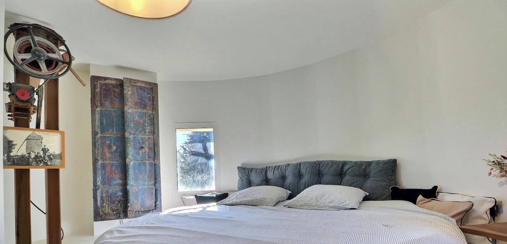 A bedroom with a large bed, a window with a view of trees, and a rustic wall with a metal door.