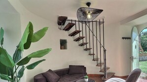 Una sala moderna con un sofá marrón, una mesa de centro redonda, una planta grande en maceta, una escalera con barandilla negra y una lámpara colgante.