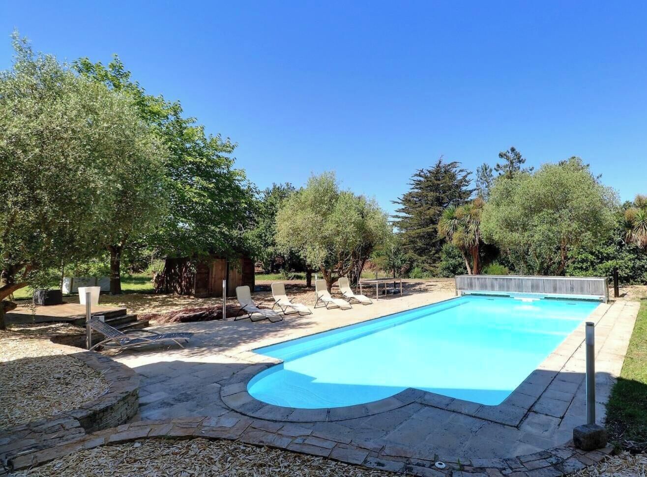 A rectangular swimming pool with a stone deck surrounding it, shaded by trees, and equipped with sun loungers.