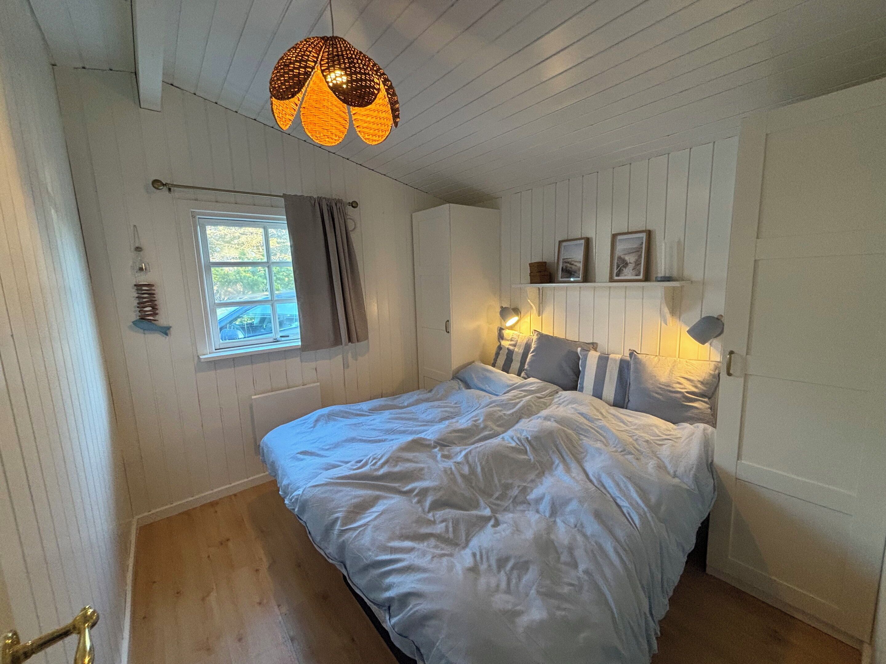 Ein Schlafzimmer mit Bett, Fenster, einer Pendelleuchte und einem an der Wand befestigten Regal mit Bildern.