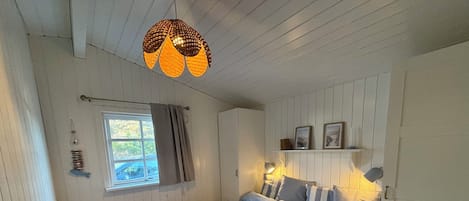 Makuuhuoneessa on sänky, ikkuna, riippuvalaisin ja seinälle kiinnitetty hylly, jossa on valokuvia.