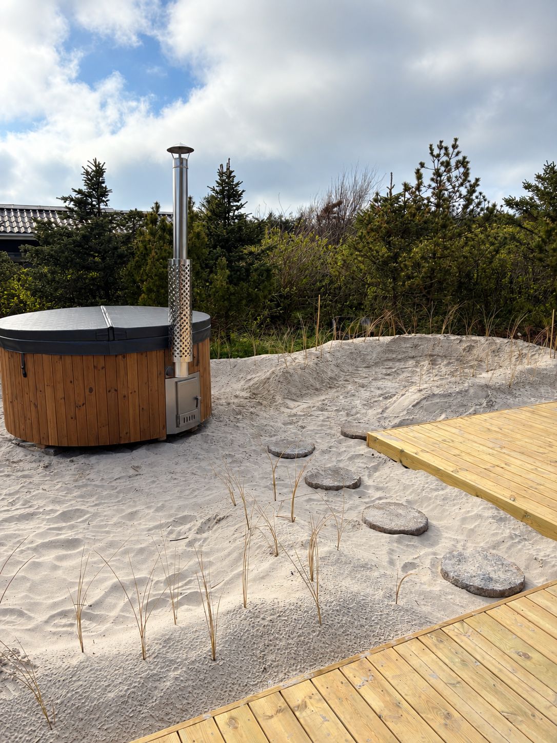Ein Whirlpool mit Holzverkleidung und einem Metallstab, umgeben von Sand und Holzstufen.