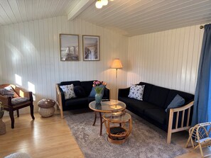 Ein Wohnzimmer mit einer Couch, Sesseln, einem Glastisch, einer Lampe und zwei gerahmten Bildern an der Wand.