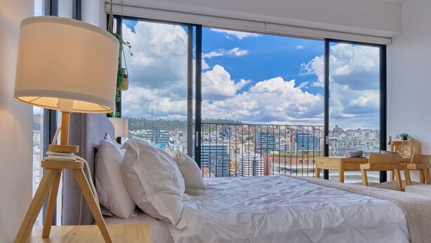 Un dormitorio con una cama grande, una mesita de noche, una lámpara y vistas a la ciudad a través de amplios ventanales.