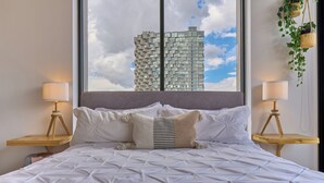 Una cama con colcha de estampado geométrico, cabecera tapizada en gris y vista a un edificio moderno a través de un ventanal grande.