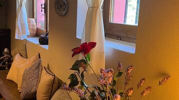 Een gezellige kamer met een houten bankje, kussens, een vaas met gedroogde bloemen en een raam met gordijnen.