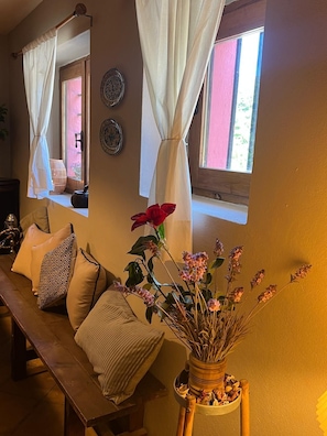 A cozy room with a wooden bench, pillows, a vase with dried flowers, and a window with curtains.