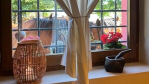 Ein Fenster mit Vorhang, ein gewebter Korb mit Blumen und ein Mörser mit Stößel auf einer Fensterbank.