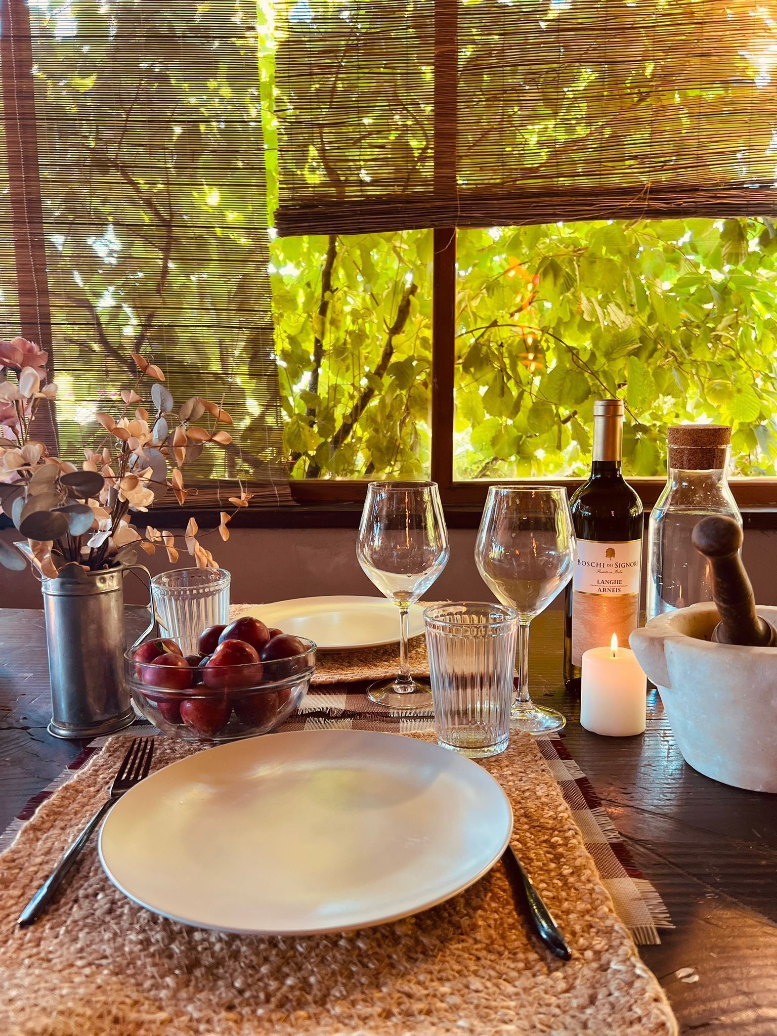 Una mesa de comedor dispuesta con platos, cubiertos, copas de vino, una vela y un frutero.