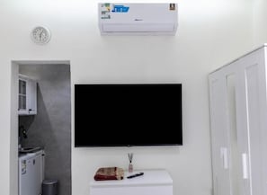 Una habitación con una televisión de pantalla plana montada en la pared, un mueble blanco pequeño, un calentador y una puerta.