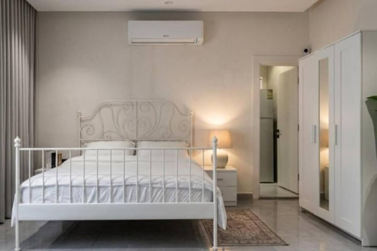 A bedroom with a white metal bed, a bedside table with a lamp, a wardrobe, and a potted plant.