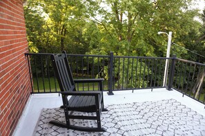 A balcony with a black rocking chair, a white patterned rug, and a view of a green park with trees.