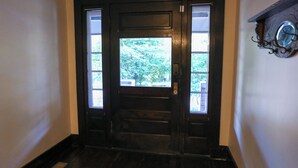 Een zwarte houten deur met een ruit en een klein raam, die uitkomt op een gang met een houten vloer en een klok aan de muur.