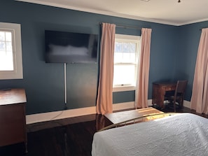 A hotel room with a bed, a desk, a chair, a flat-screen TV mounted on the wall, and a window with curtains.