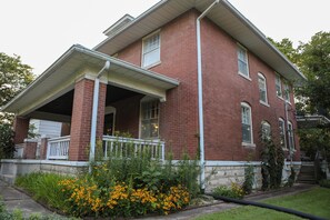 A two-story brick house with a porch and a small garden with yellow flowers.