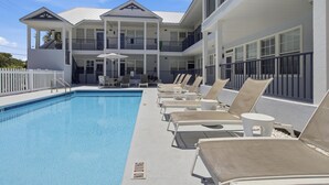 Une piscine avec des chaises longues et des parasols, une clôture blanche et un bâtiment doté de balcons.