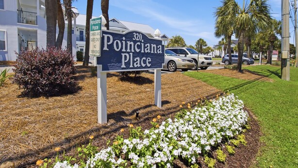 Un panneau indiquant « Poinciana Place », offrant une vue dégagée sur les environs, notamment un bâtiment, des voitures garées et un jardin aménagé.