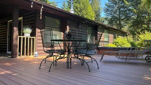 Um deck de madeira com mesa e cadeiras de metal, rodeado por uma cabana com janelas amplas e uma chaminé.