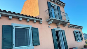 Un immeuble de deux étages avec des volets verts, un balcon orné de balustrades en fer forgé et un escalier en pierre menant à l’entrée.