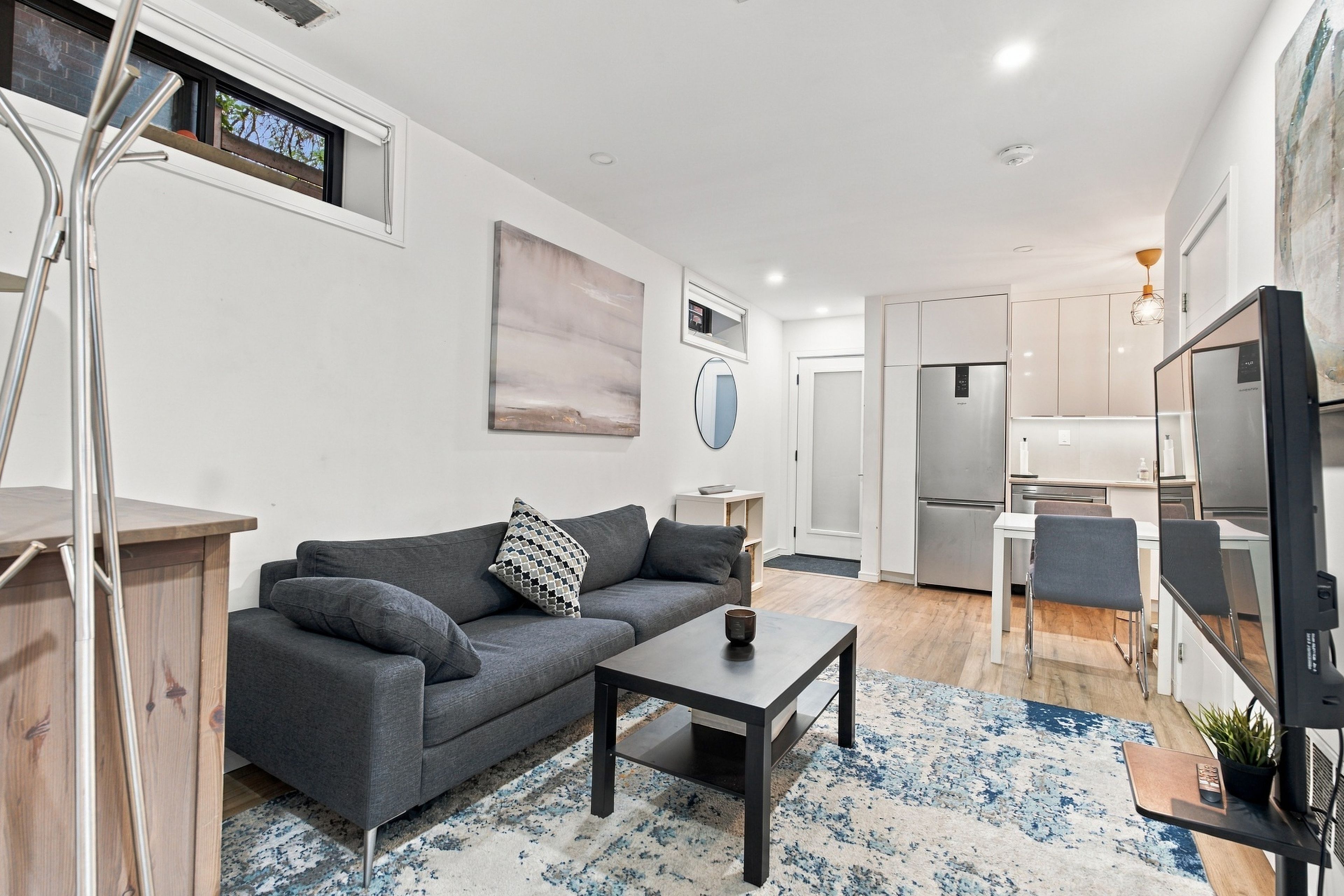 Un espacio habitable moderno con un sofá gris, una mesa de centro, un televisor de pantalla plana y una zona de cocina con un refrigerador.