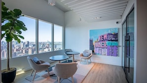 A modern office space with large windows, a city view, a potted plant, and a colorful artwork on the wall. - Luxury 1BR, Skyline Views, Penn Station (New York)