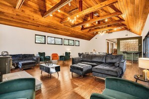 Ein großzügiger Wohnbereich mit hoher Holzbalkendecke, verschiedenen Sitzgelegenheiten wie Sofas und Sesseln sowie großen Fenstern für natürliches Licht.