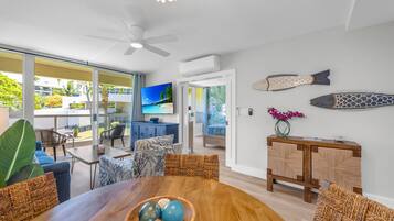 A modern living room with a wooden dining table, wicker chairs, a TV, and a balcony with a view of the ocean.