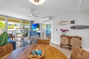 A modern living room with a wooden dining table, wicker chairs, a TV, and a balcony with a view of the ocean. - New South Kihei Beach Resort Vistas (Kihei)